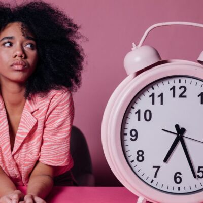 Pensive woman with afro hair leaning on a table next to a large pink alarm clock.