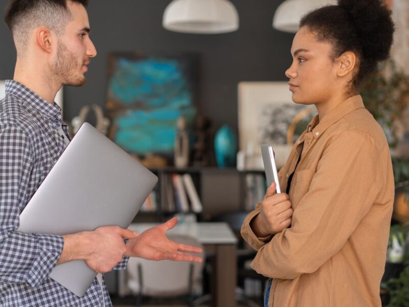 Two office workers engaged in a conversation, holding devices, in a modern office setting.
