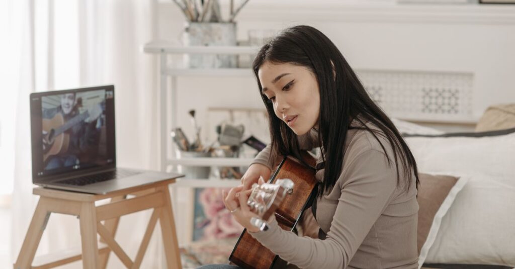Como a Prática de Hobbies Pode Aumentar sua Autoestima 2 Asian woman taking an online guitar lesson at home using a laptop.
