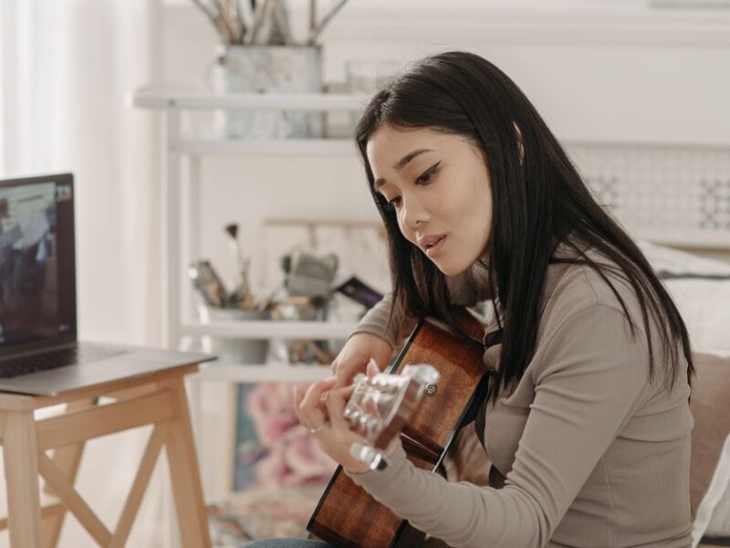 Asian woman taking an online guitar lesson at home using a laptop.