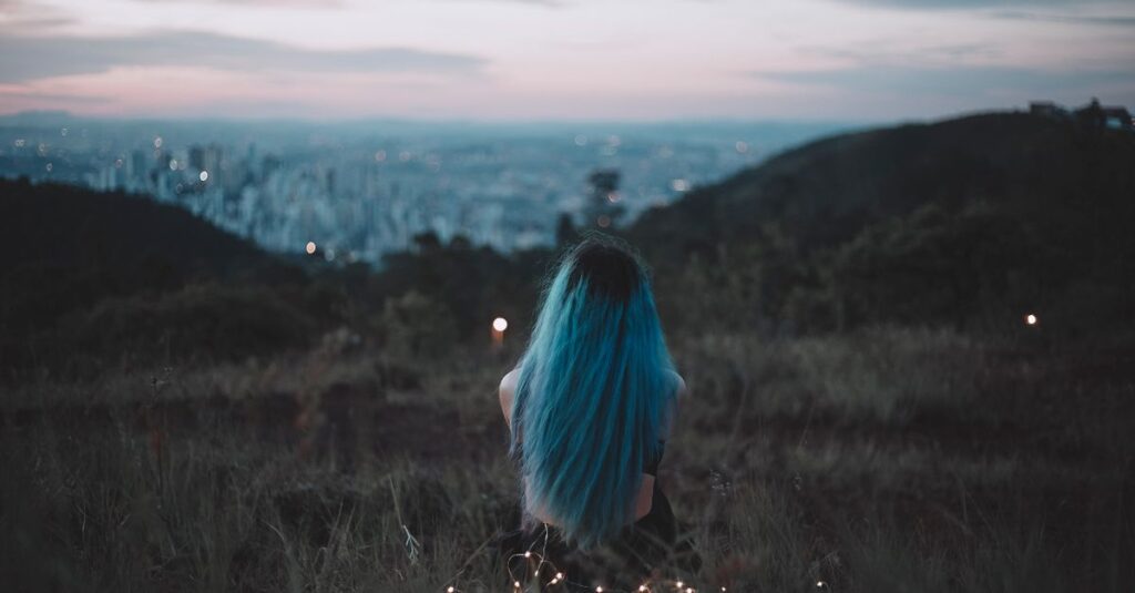 A woman with blue hair sits on a hillside, gazing at the city's twilight skyline adorned with string lights.