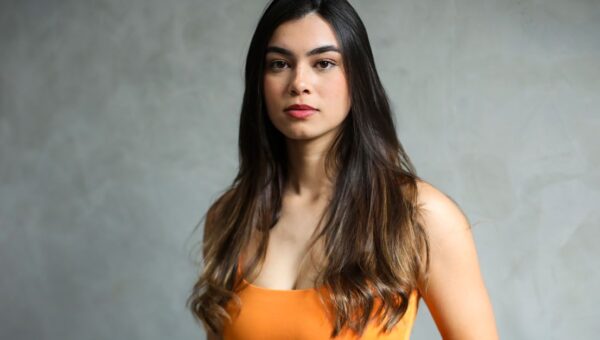 Elegant portrait of a young woman with long, dark hair in a vibrant orange top against a neutral background.