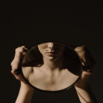 Artistic portrait of a woman holding a mirror, reflecting her face, with a dark, moody background.