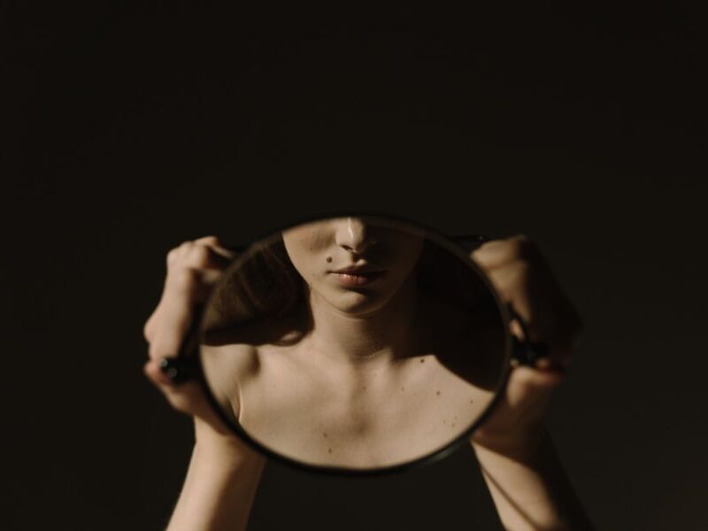 Artistic portrait of a woman holding a mirror, reflecting her face, with a dark, moody background.